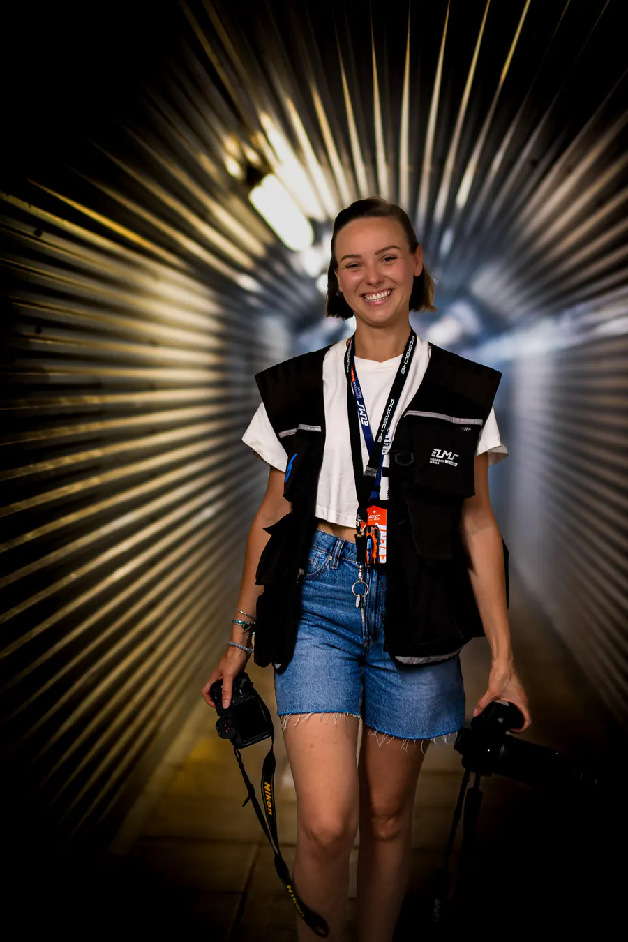 Diana Zayats sorride nel tunnel dei box con due macchine fotografiche in mano e il gilet da fotografa accreditata