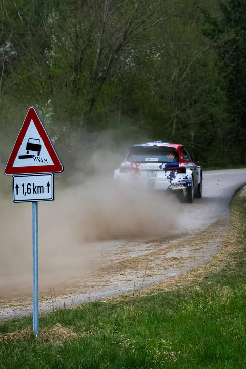 Vettura da rally solleva polvere su una prova speciale sterrata accanto a un cartello stradale